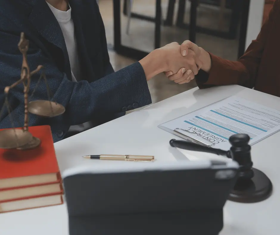 Two individuals shaking hands at a desk, surrounded by legal papers and a scale, symbolizing successful mediation.